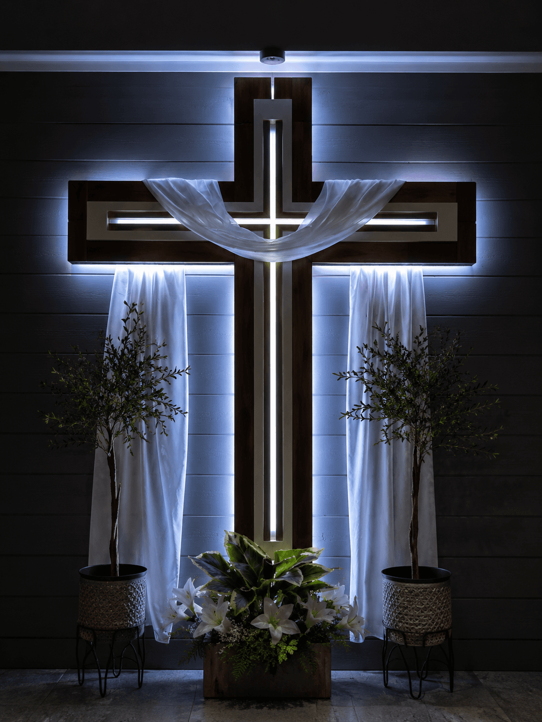 High-resolution 4K image of a modern illuminated wooden Christian cross draped with a white cloth, decorated with flowers and plants, ideal for church interiors, Easter displays, and Christian wallpapers.